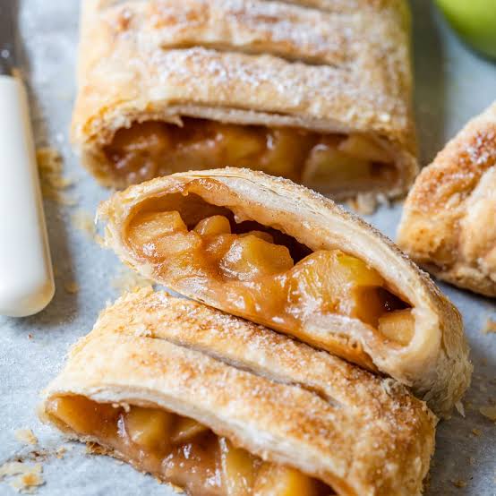 An apple strudel cut into portions, its puff pastry dusted with icing sugar and chunky apple filling showing